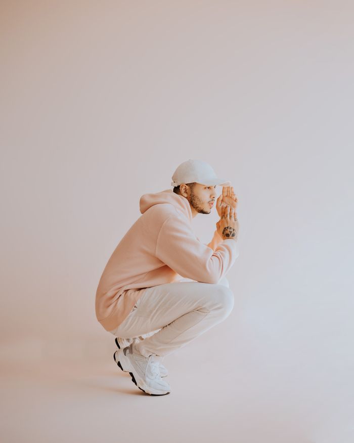 gallery-3 Young man with tattoos in pastel attire squats elegantly, showcasing street style against a white background.